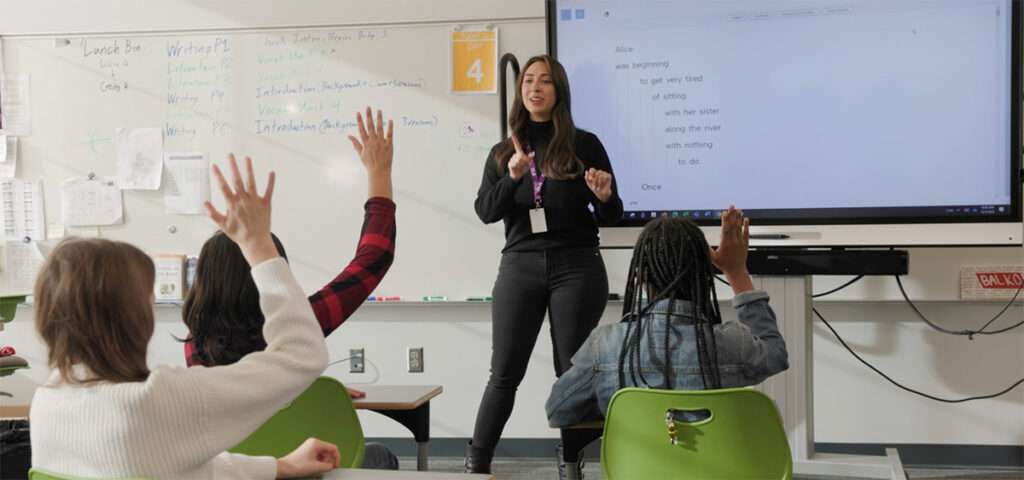 Teacher standing at the front of a classroom near a large screen displaying cascading text. Three students, seated at their desks with raised hands, are waiting to be called on by the teacher.